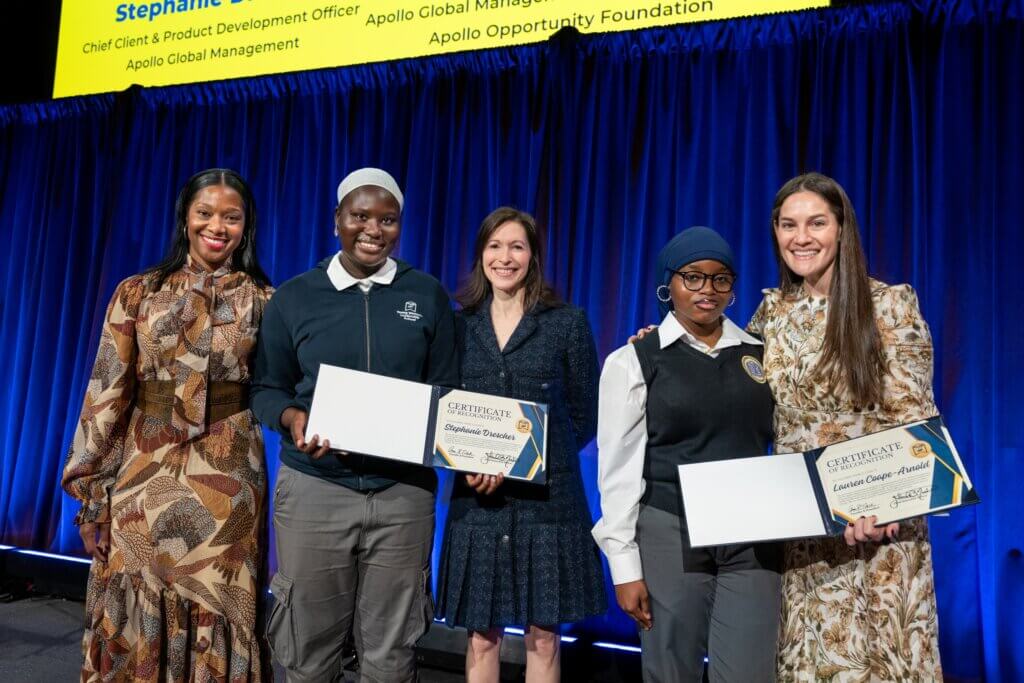Five women stand in front of a blue curtain at an event, three of whom are holding certificates and smiling at the camera.