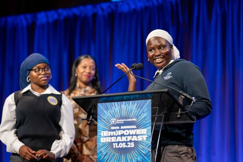 Three people stand on stage at a podium, with one student wearing her school uniform and a hijab speaking and gesturing while the others look on.