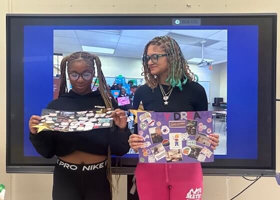 Two students stand in front of a large screen, each holding a decorated poster board with various images and text.