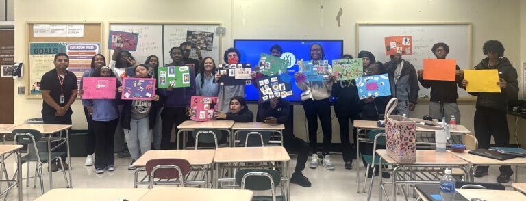 A group of students stands in a classroom holding up colorful project posters