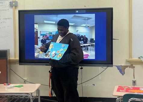 A student stands in front of a screen displaying a classroom photo, holding and presenting a colorful poster board to the class.