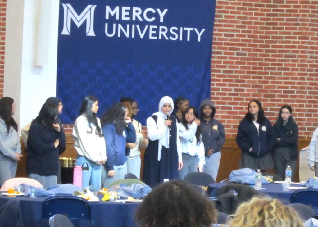 A group of students stand in front of a Mercy University banner; one student speaks into a microphone