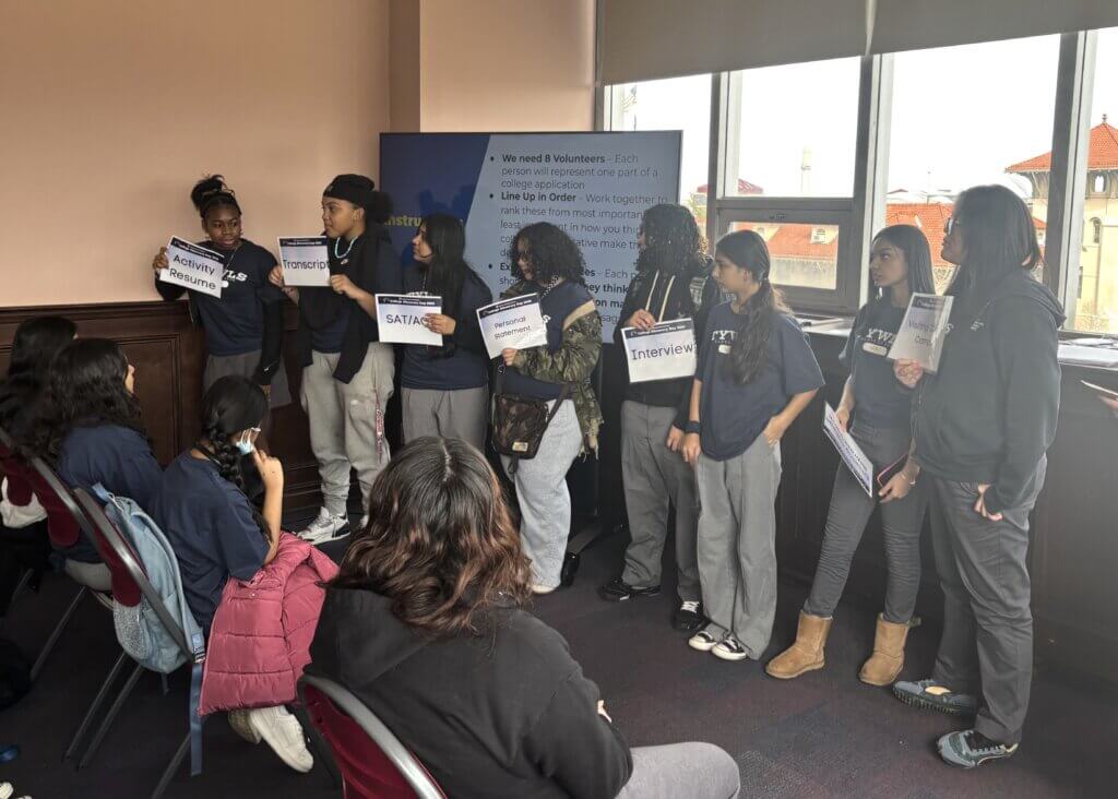 Students in a Girls' Education program stand holding signs labeled Activity Presenter, SAT/ACT, Transcript, and Interview, while others sit and listen.