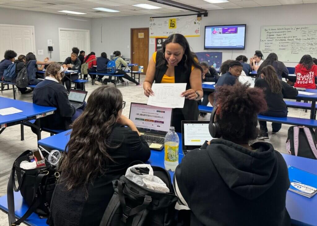 A teacher distributes papers to students working on laptops at blue tables in a classroom, with more students visible in the background.