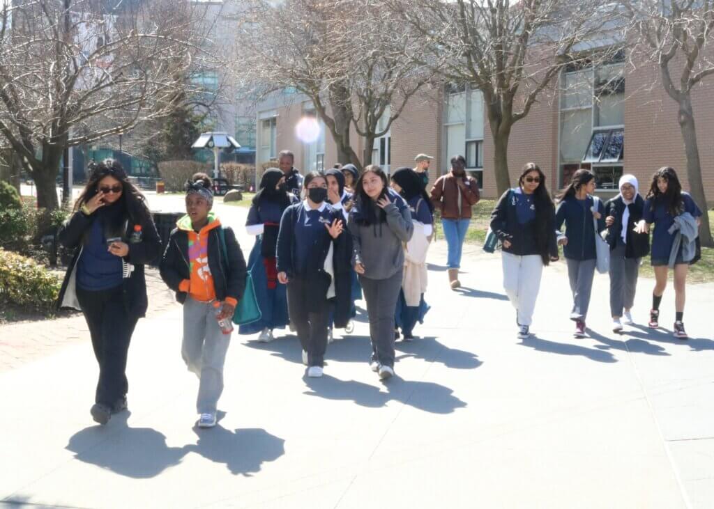 A group of students and adults walk together outside by a tree-lined path on the Queens College campus