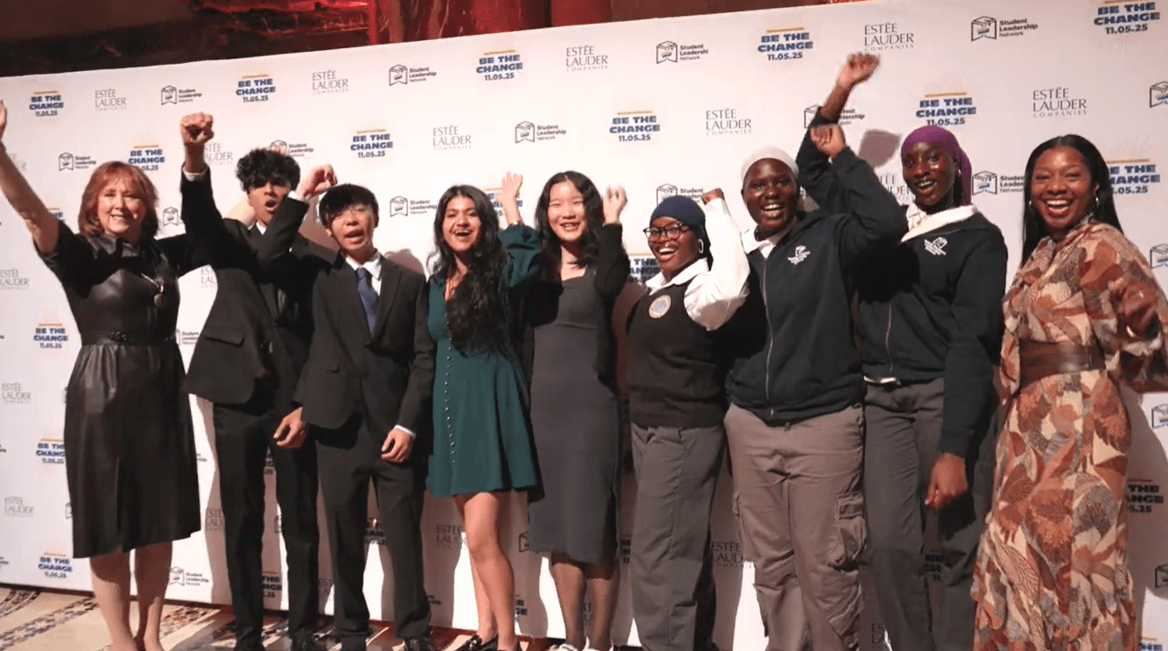 A group of nine people stand in front of a branded EmPower Breakfast event backdrop, smiling and raising their hands in celebration