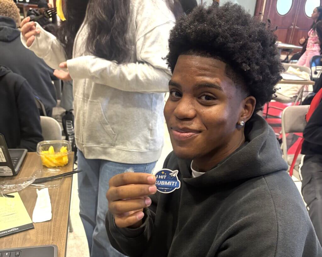 A smiling student sitting at a table holds a sticker that reads I HIT SUBMIT! in a classroom setting.