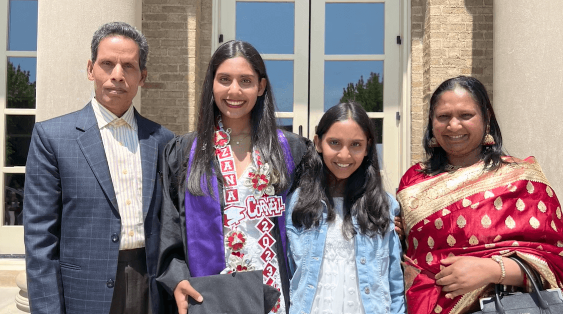 Four family members stand together outside a building. One woman wears a graduation gown and sash from the Young Women's Leadership School of Queens.