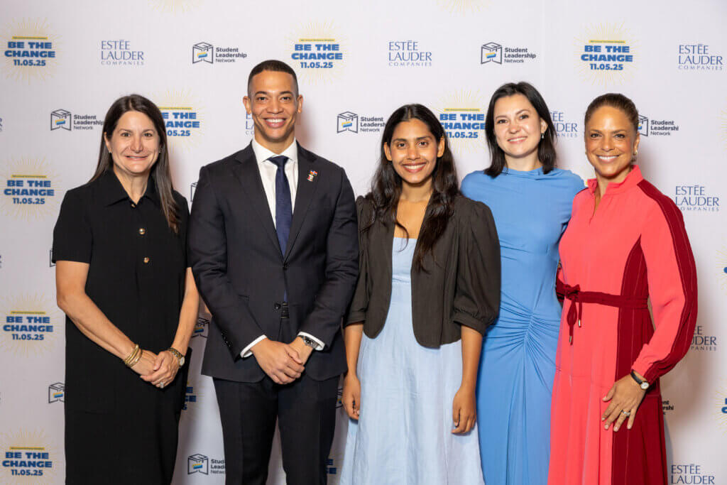 Five people stand together, smiling for a group photo in front of a backdrop featuring Be the Change, Estée Lauder Companies, and Student Leadership Network logos.