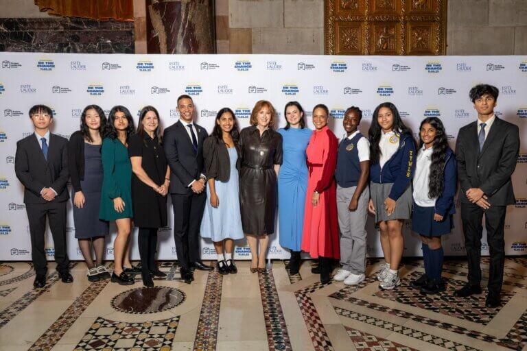 A group of people, including adults in formal attire and students in uniforms, pose together in front of a step-and-repeat event banner indoors.
