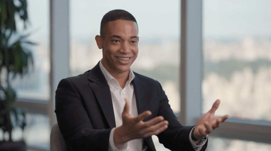 A 2025 Power Breakfast Honoree sits indoors in a suit, smiling and gesturing with both hands, framed by large windows