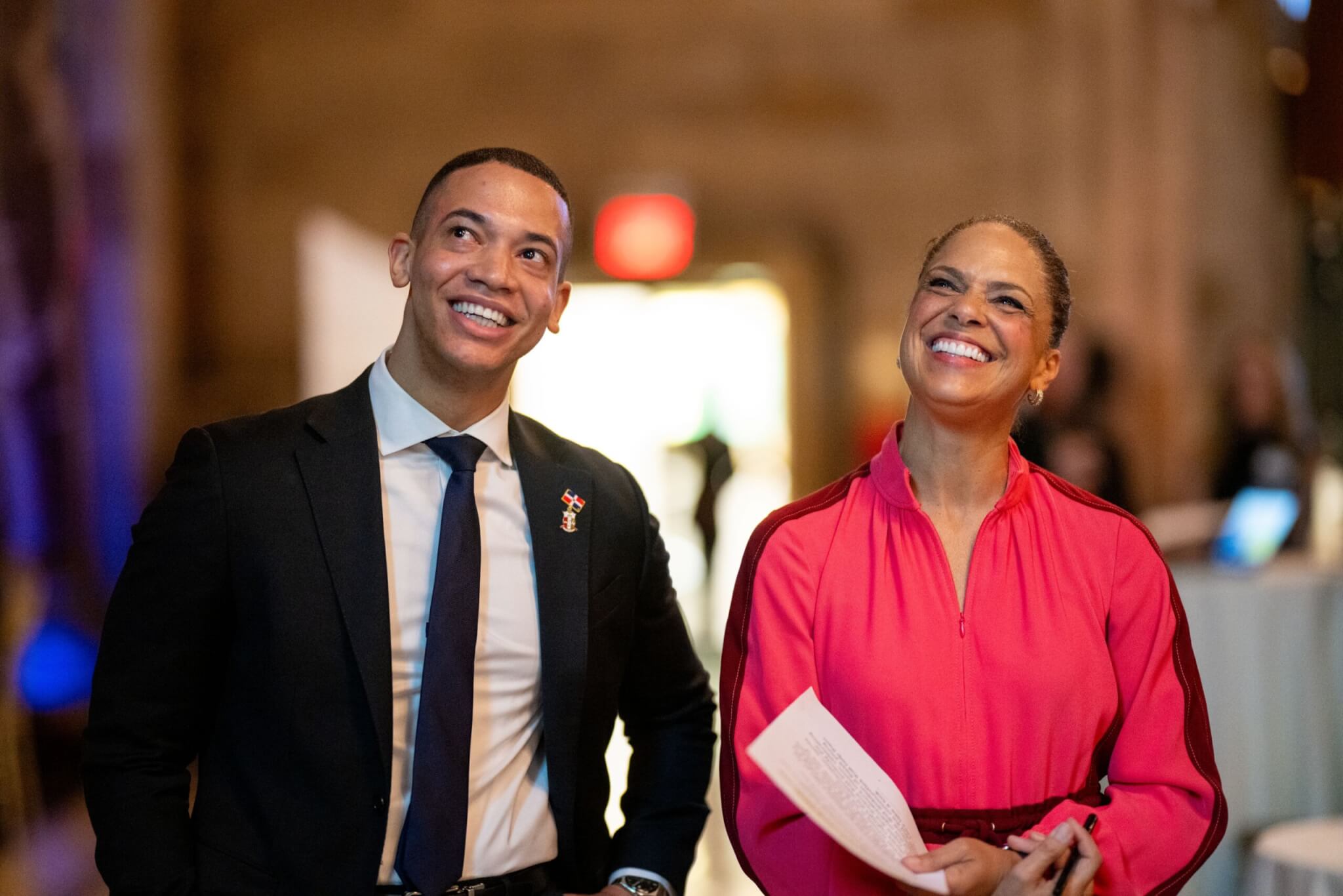 Two people in formal attire stand indoors, smiling and looking upward; one holds a piece of paper. The background is softly blurred.