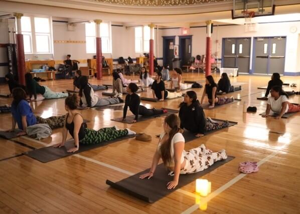 A group of students practice yoga on mats in a gymnasium, performing upward-facing poses. A lit candle is placed on the floor in the foreground.