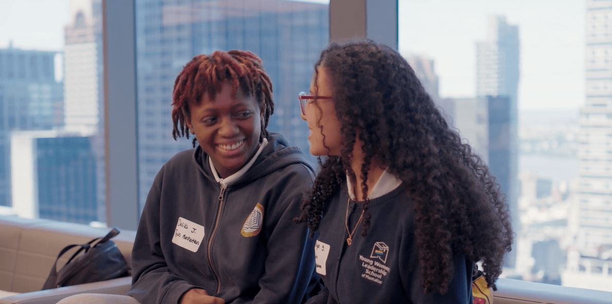 Two young people wearing name tags and sweatshirts sit indoors by a large window with city buildings in the background, smiling and talking during the Apollo Opportunity Foundation (Em)Power Breakfast.