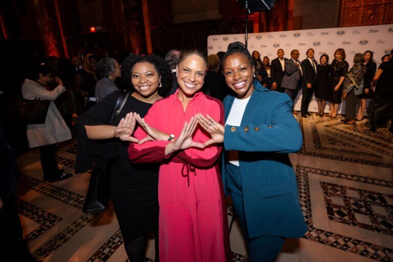 Three women stand together at an indoor event, smiling and making a triangular hand gesture representing their soririty