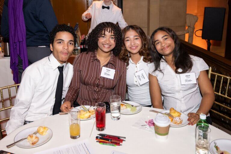 Four students sit at a round table with food and drinks, smiling at the camera