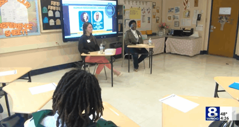 Two speakers sit at desks in front of a classroom, addressing students seated at desks.
