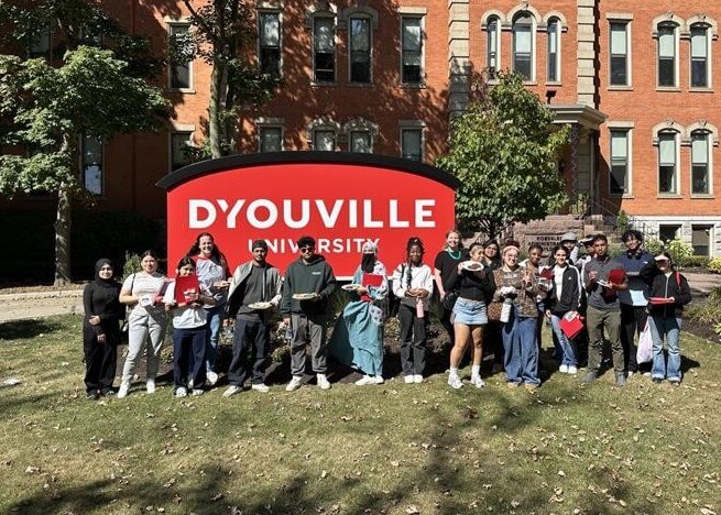 A group of students stands in front of a large red DYouville University sign on a lawn with a brick building in the background.