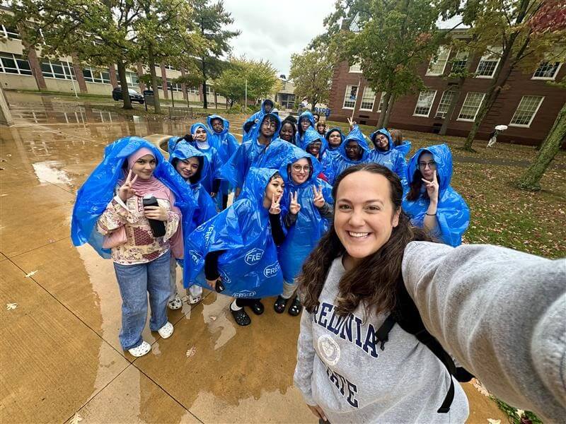 A group of people in blue rain ponchos pose for a selfie outdoors on a rainy day near a school building on the SUNY Fredonia campus