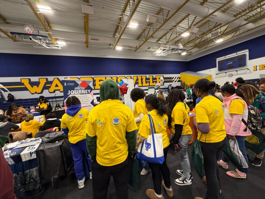 A group of people in yellow shirts gather around booths at an indoor HBCU college fair visit