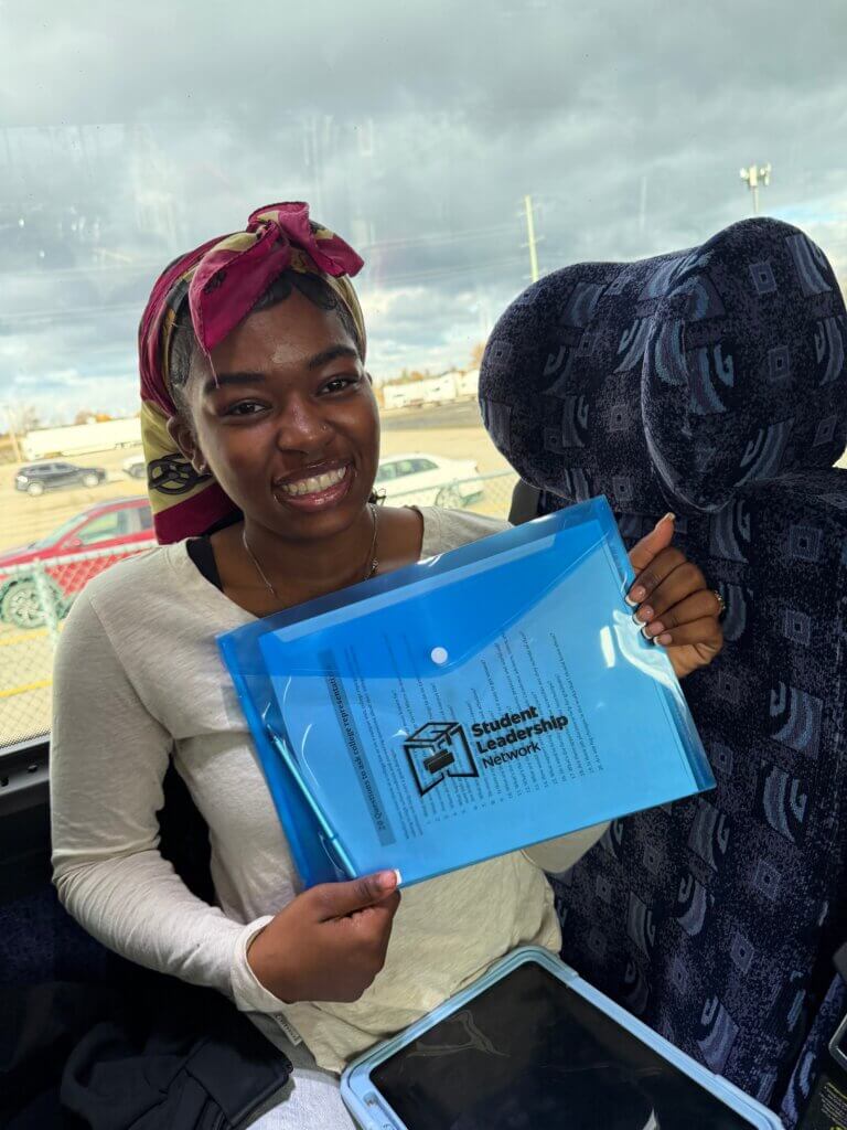 A person sitting on a bus seat smiles while holding a blue folder labeled Student Leadership Network.