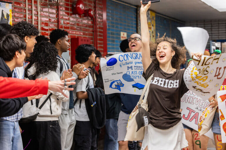 A group of students cheer in a hallway as one student holding a college poster raises her arm in celebration, wearing a Lehigh t-shirt.