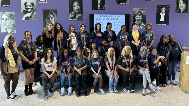 A group of students wearing college shirts pose for a photo in front of a wall with historical portraits of women trailblazers behind them.