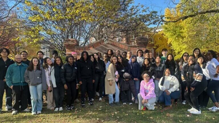 A group of people pose together outdoors in front of a brick structure at Gettysburg College, surrounded by trees with autumn foliage on a sunny day.