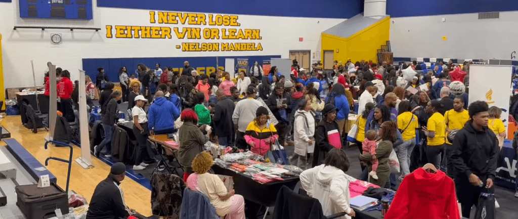 Large crowd gathers in a gymnasium for an indoor HBCU college fair visit with tables, merchandise, and a Nelson Mandela quote displayed on the wall.