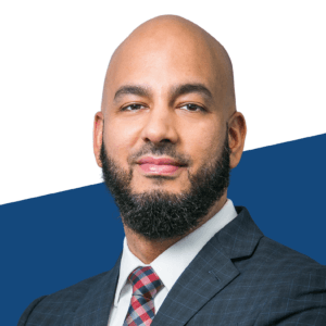 A bald man with a trimmed beard, dressed in a checkered suit, white shirt, and patterned tie, stands against a white and blue background—perfect for a higher education podcast or an RBC Capital Markets feature.