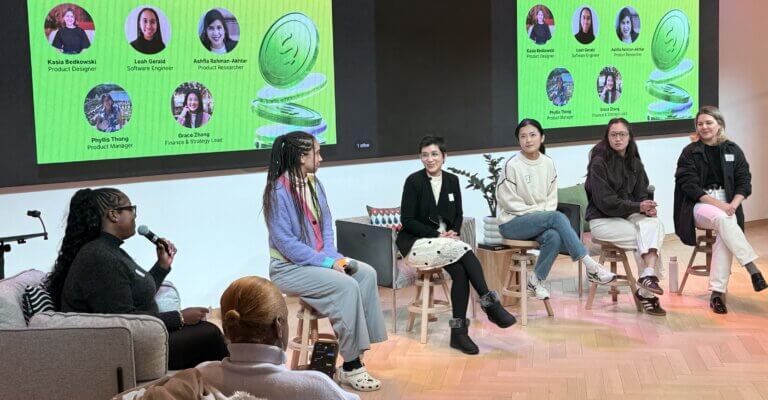 Six women sit on stools in front of a screen displaying their names and roles, participating in a panel discussion