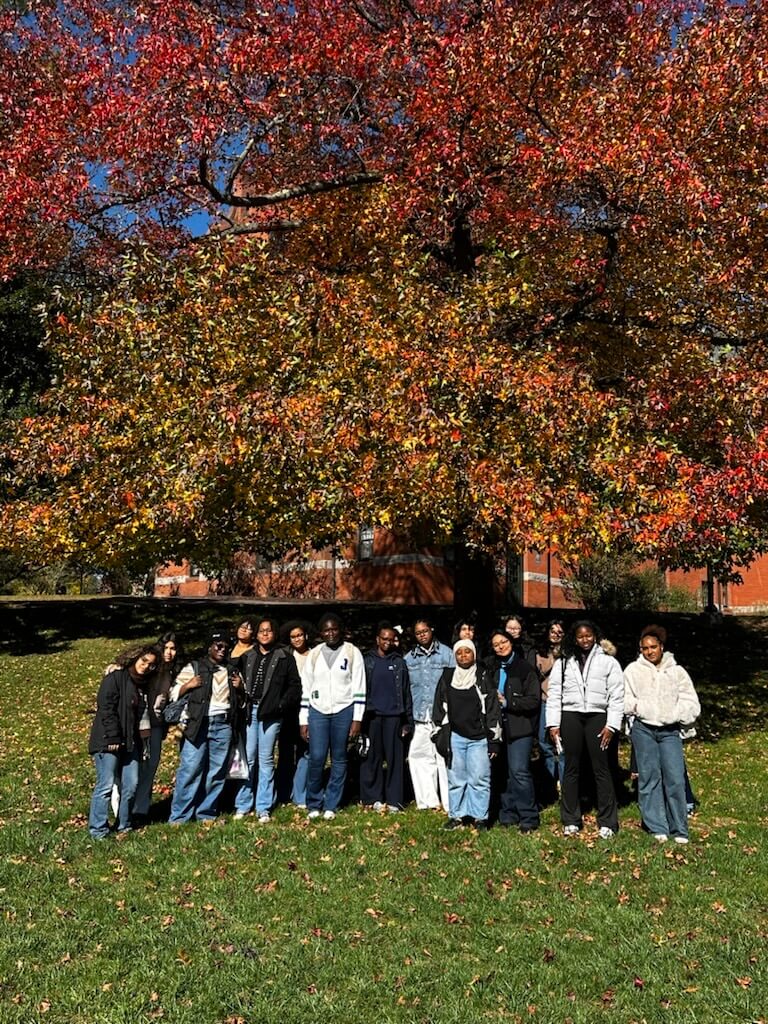 High school students and counselors stand in front of a large tree with red and orange autumn leaves on the Mount Holyoke College campus.