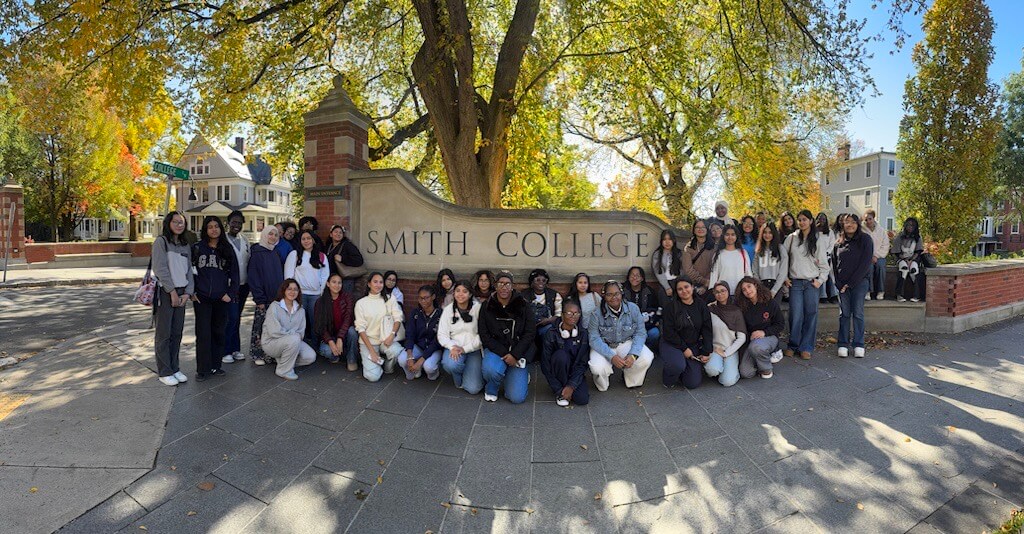 High school students and counselors pose in front of the Smith College entrance sign on a sunny day surrounded by trees and buildings.
