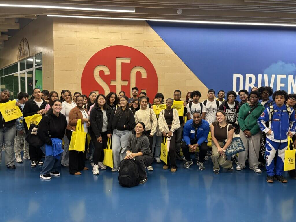 A large group of students pose together indoors in front of an SFC logo, holding yellow tote bags and smiling at the camera.