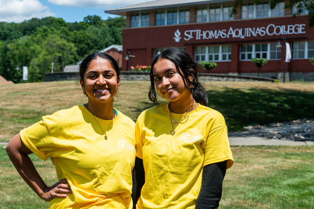 Two young women in yellow shirts stand outside in front of a building labeled St. Thomas Aquinas College.