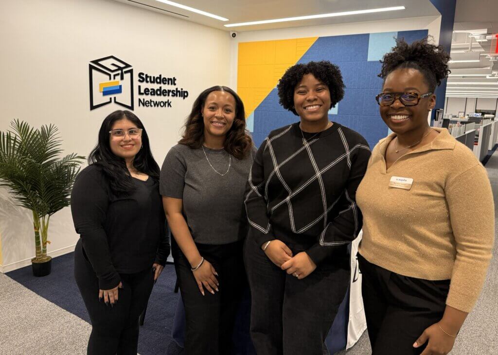 Four alumna stand and smile in an office with a Student Leadership Network sign on the wall behind them.