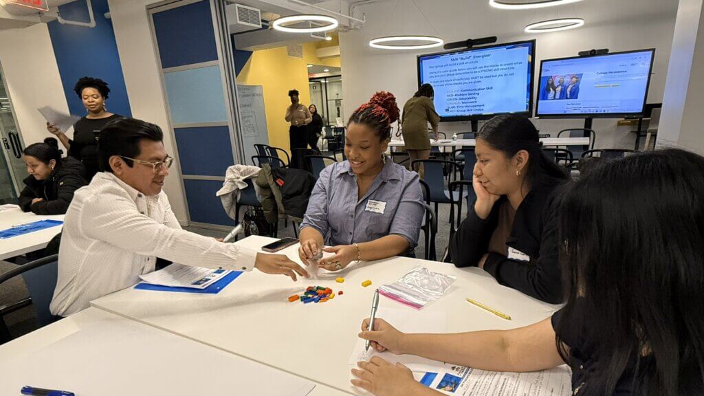 Four people sit at a table in a classroom, interacting with colorful Lego blocks and taking notes, while others work in the background.