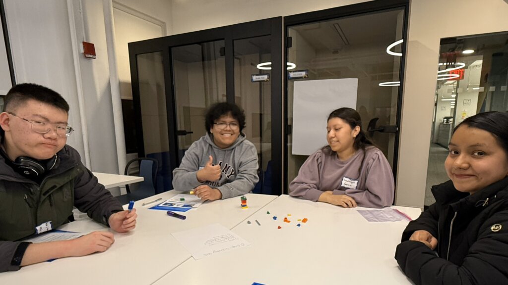 Four alumni and students sit around a table with papers and small objects, engaging in an activity; one person is giving a thumbs up