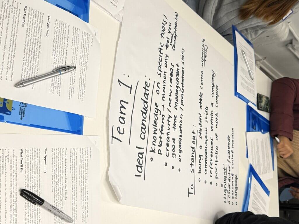 A large worksheet titled Team 1: Ideal candidate lists desired qualities and ways to stand out, placed on a table with pens, documents, and people seated around it.