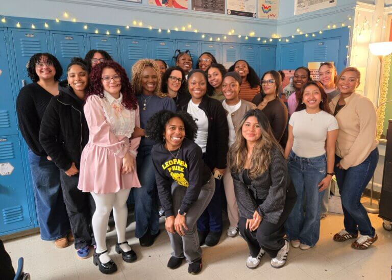 A group of young adults stands and kneels together, smiling for a photo in front of blue lockers during Alumni Day to give advice about college decisions