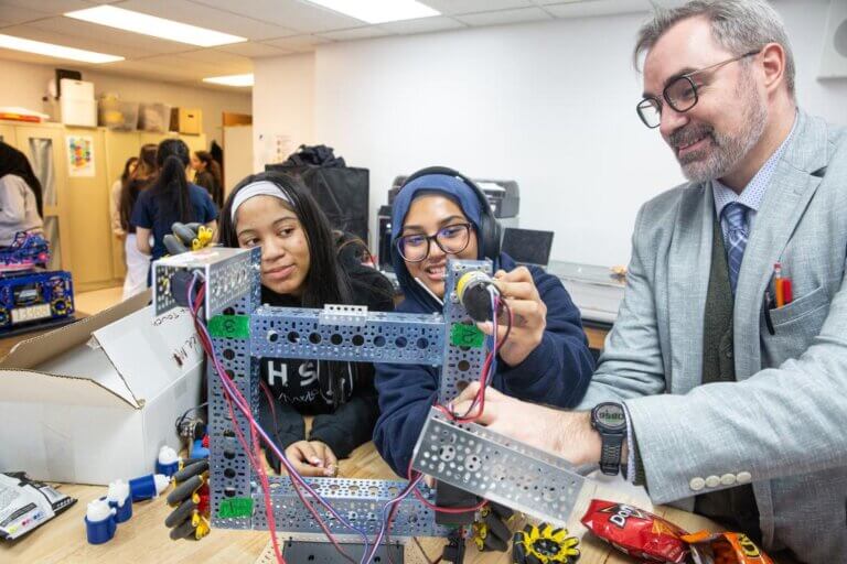 Two students and a teacher at TYWLS Astoria work together on a robotics project in a classroom, assembling metal and electronic components at a table.