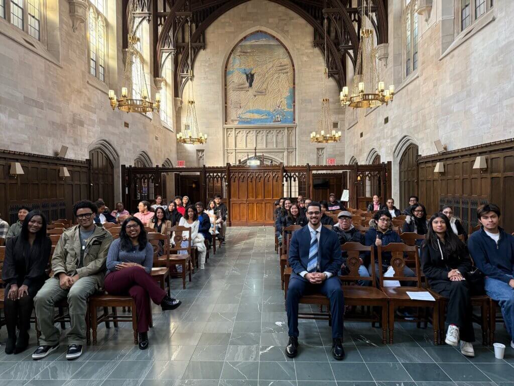 A group of people sits in rows of wooden chairs inside a large, ornate hall with high ceilings, arched windows, and a mural on the back wall.