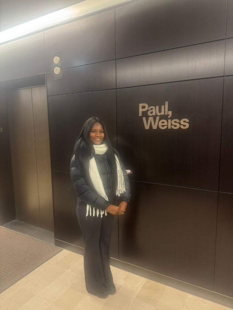 A woman in a black jacket and white scarf stands in front of a wall with the sign Paul, Weiss.