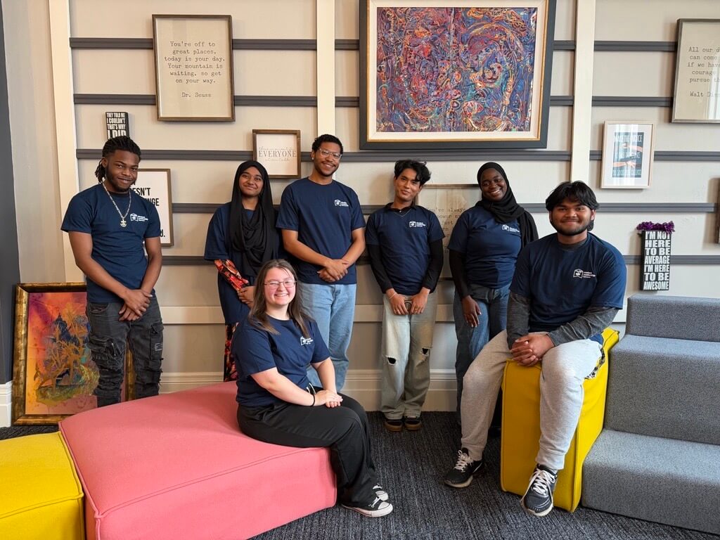 Seven college transition coaches wearing matching navy blue shirts pose together in a modern room with colorful artwork and motivational quotes on the walls.