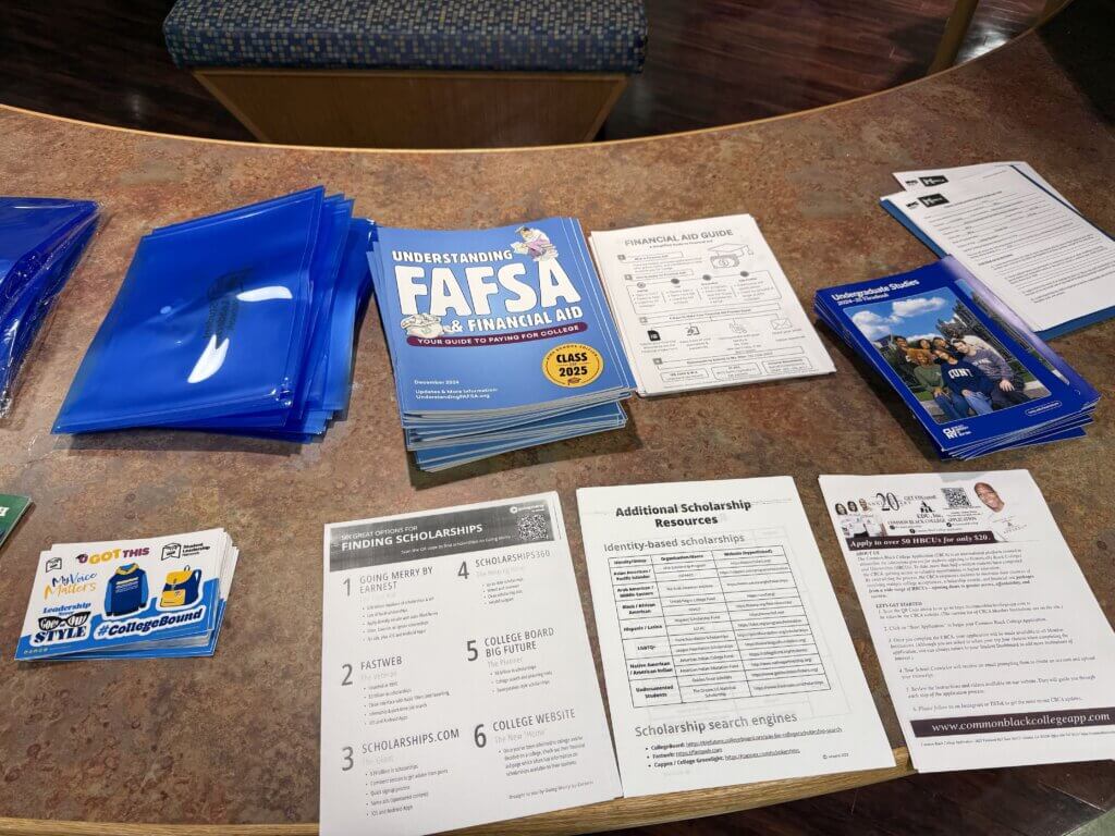 A table displays FAFSA financial aid booklets, scholarship information sheets, blue plastic folders, and informational flyers