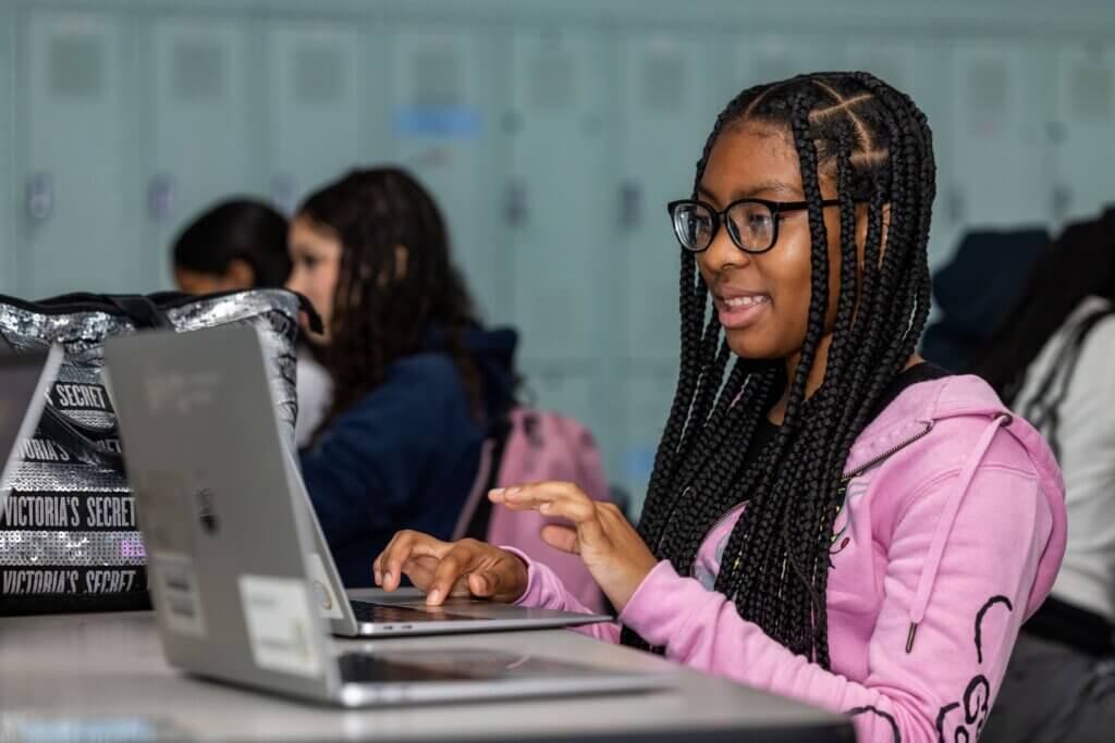 A student with braided hair and glasses types on a laptop at a desk in a classroom