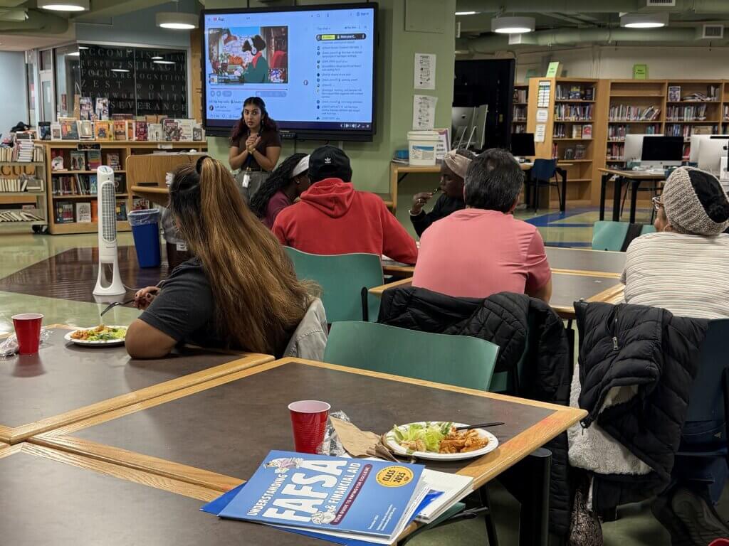 Families sit at tables while a person stands near a screen displaying a presentation, ready to provide college counseling for families. A FAFSA booklet is on a table in the foreground.