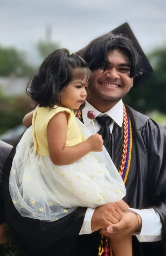 A young man in a graduation cap and gown smiles while holding his younger sister who is in a yellow and white dress