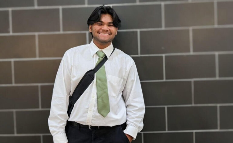 A young man wearing a white shirt, light green tie, and black shoulder bag enters a school building