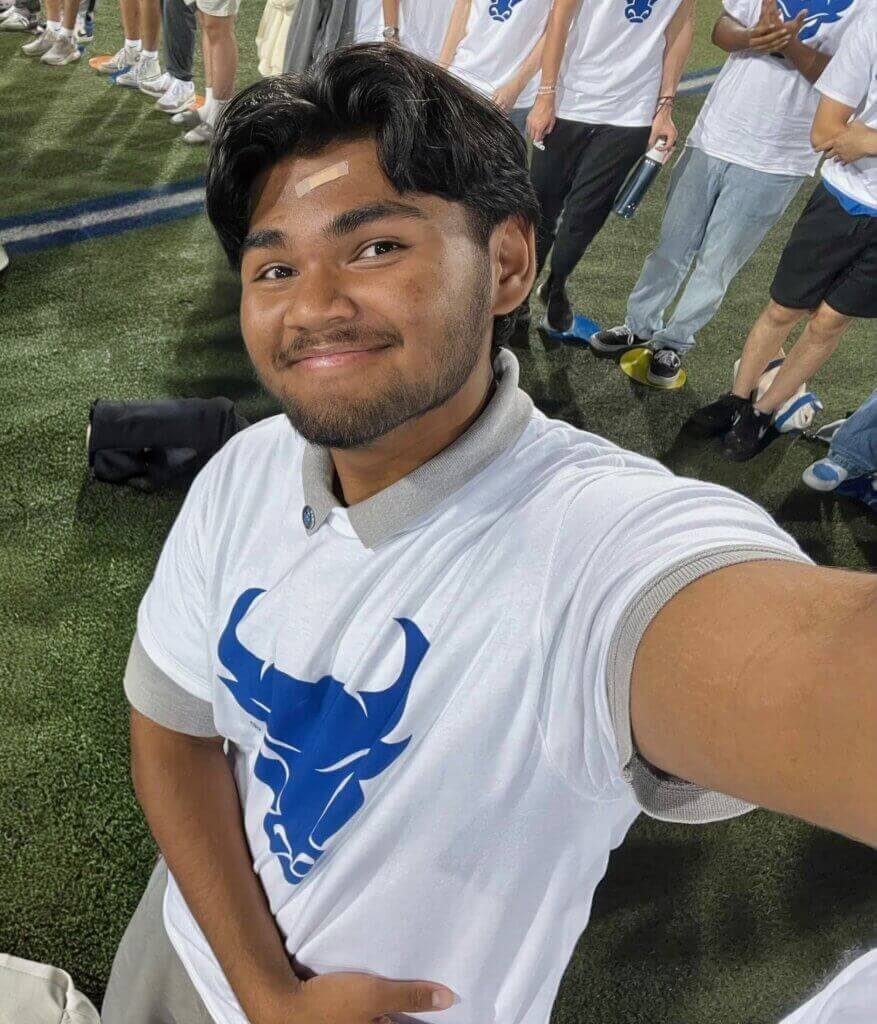 A young man wearing a white shirt with a University at Buffalo blue bull logo takes a selfie on a sports field, surrounded by others in similar shirts.